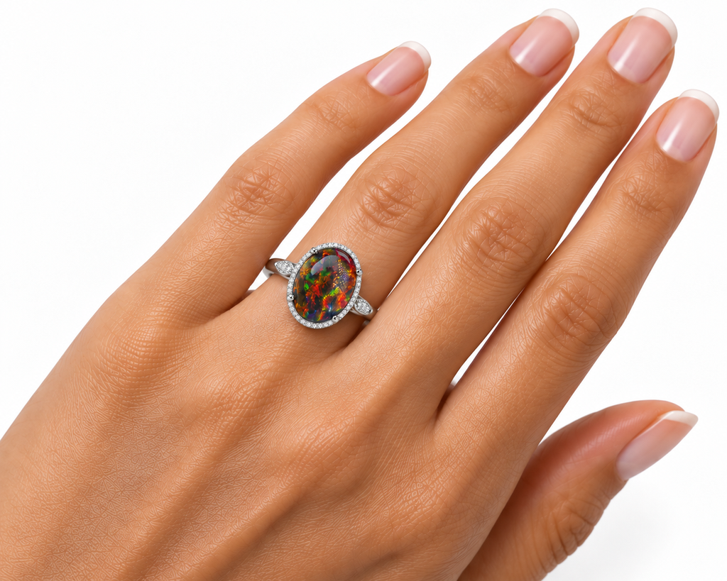 A close-up of a hand wearing a silver-toned ring with a round iridescent opal center stone and a halo of small clear stones, set against a plain white background.