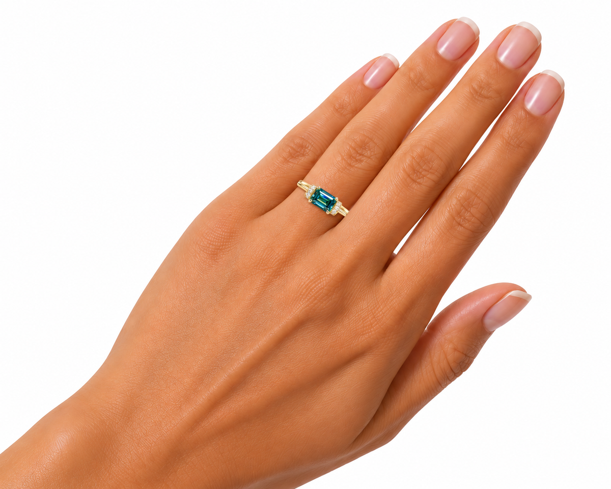 Angled close-up of a woman's hand wearing a 1 carat green moissanite ring, showcasing the elongated 5x7mm east-west emerald-cut gemstone, polished gold-tone four-prong setting, and delicate cubic zirconia shoulder accents against a white background.