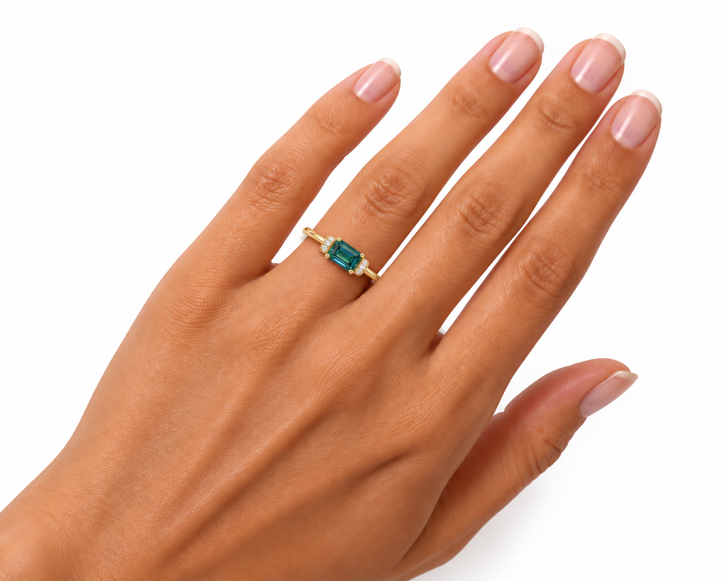 1 carat lab-created green moissanite ring worn naturally on a woman's ring finger, featuring a true-to-size 5x7mm horizontally positioned emerald-cut gemstone with teal-green reflections, sparkling cubic zirconia accents, and a Gold Plated 925 sterling silver band.
