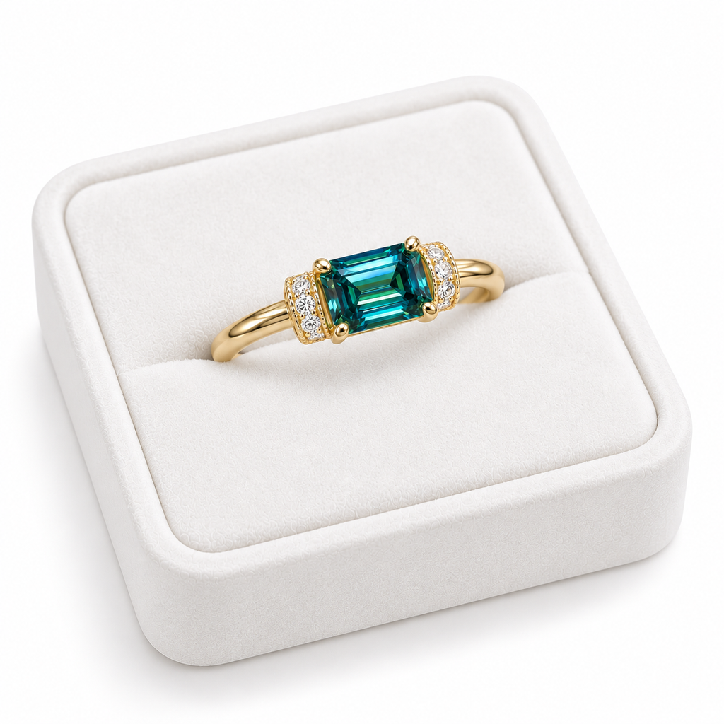 Angled view of a 1 carat green moissanite ring resting on a white velvet jewelry tray, showcasing the elongated 5x7mm emerald-cut gemstone, polished Gold Plated sterling silver band, and decorative cubic zirconia shoulder accents.