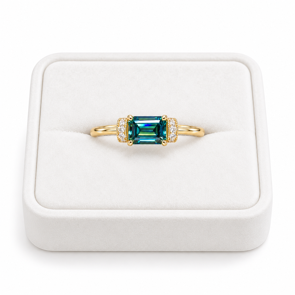 1 carat lab-created green moissanite ring featuring a horizontal 5x7mm emerald-cut gemstone with vivid teal-green reflections, four gold-tone prongs, and sparkling cubic zirconia accents, displayed on a white velvet jewelry tray against a pure white background.
