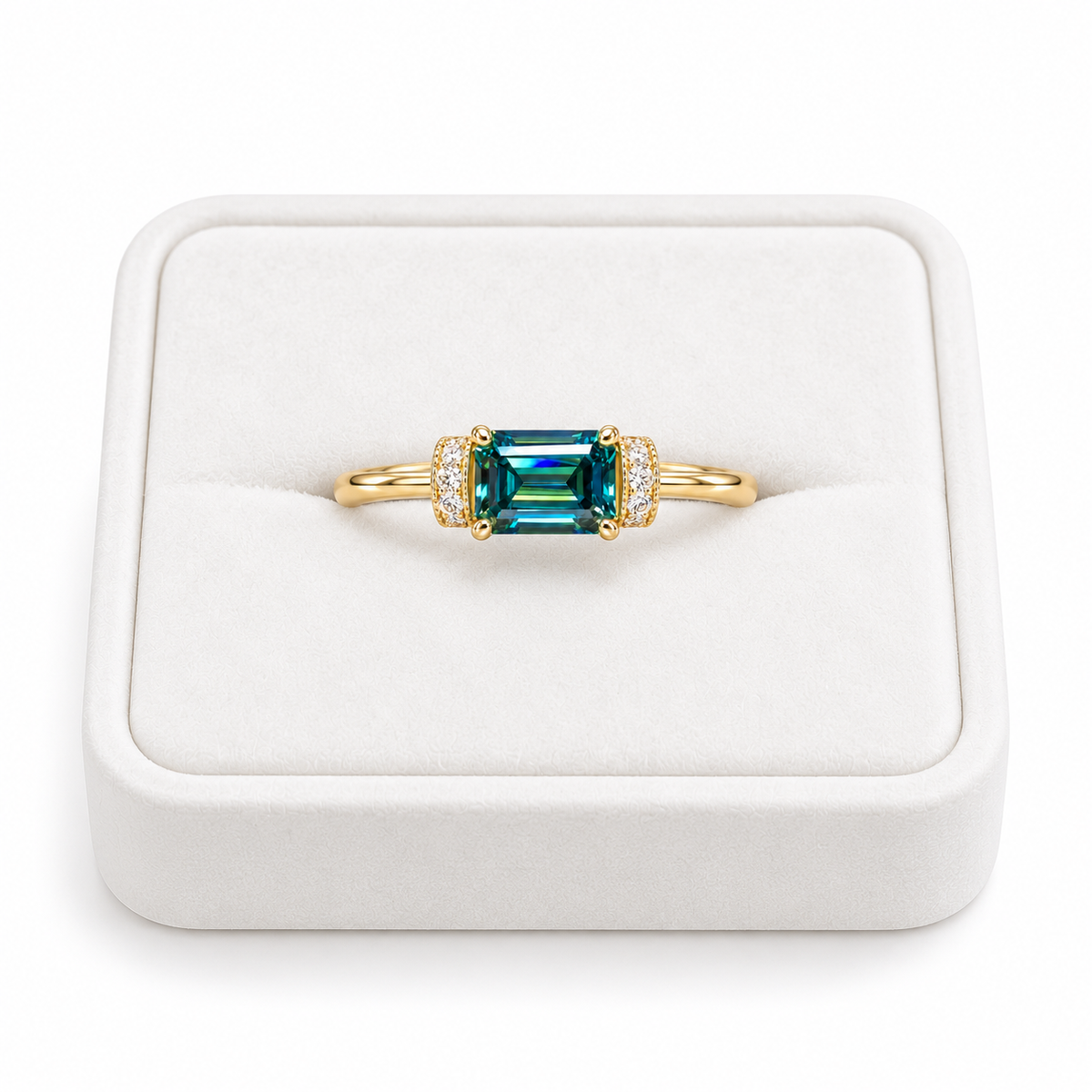 1 carat lab-created green moissanite ring featuring a horizontal 5x7mm emerald-cut gemstone with vivid teal-green reflections, four gold-tone prongs, and sparkling cubic zirconia accents, displayed on a white velvet jewelry tray against a pure white background.