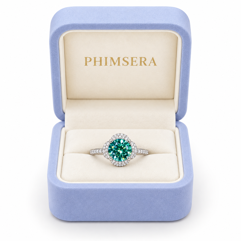 2 carat 8mm round brilliant green moissanite floral halo ring displayed inside a periwinkle Phimsera jewelry gift box with an ivory interior and gold PHIMSERA lettering, showcasing the vibrant teal-green gemstone, sparkling cubic zirconia floral halo, and polished sterling silver band against a white background.
