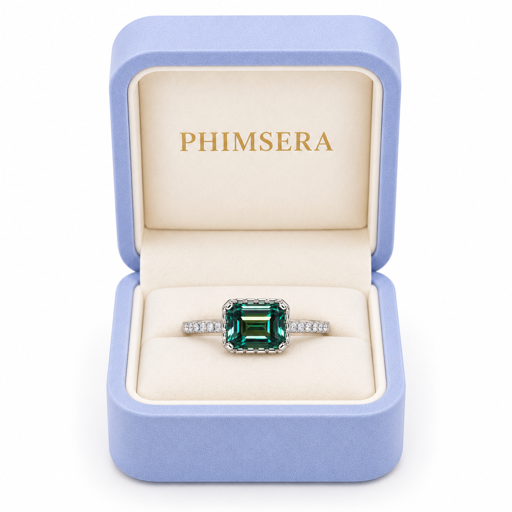 2 carat lab-created green moissanite ring displayed inside a periwinkle Phimsera jewelry box with cream velvet interior and gold PHIMSERA lettering, featuring a 6x8mm emerald-cut gemstone and sparkling cubic zirconia accents.