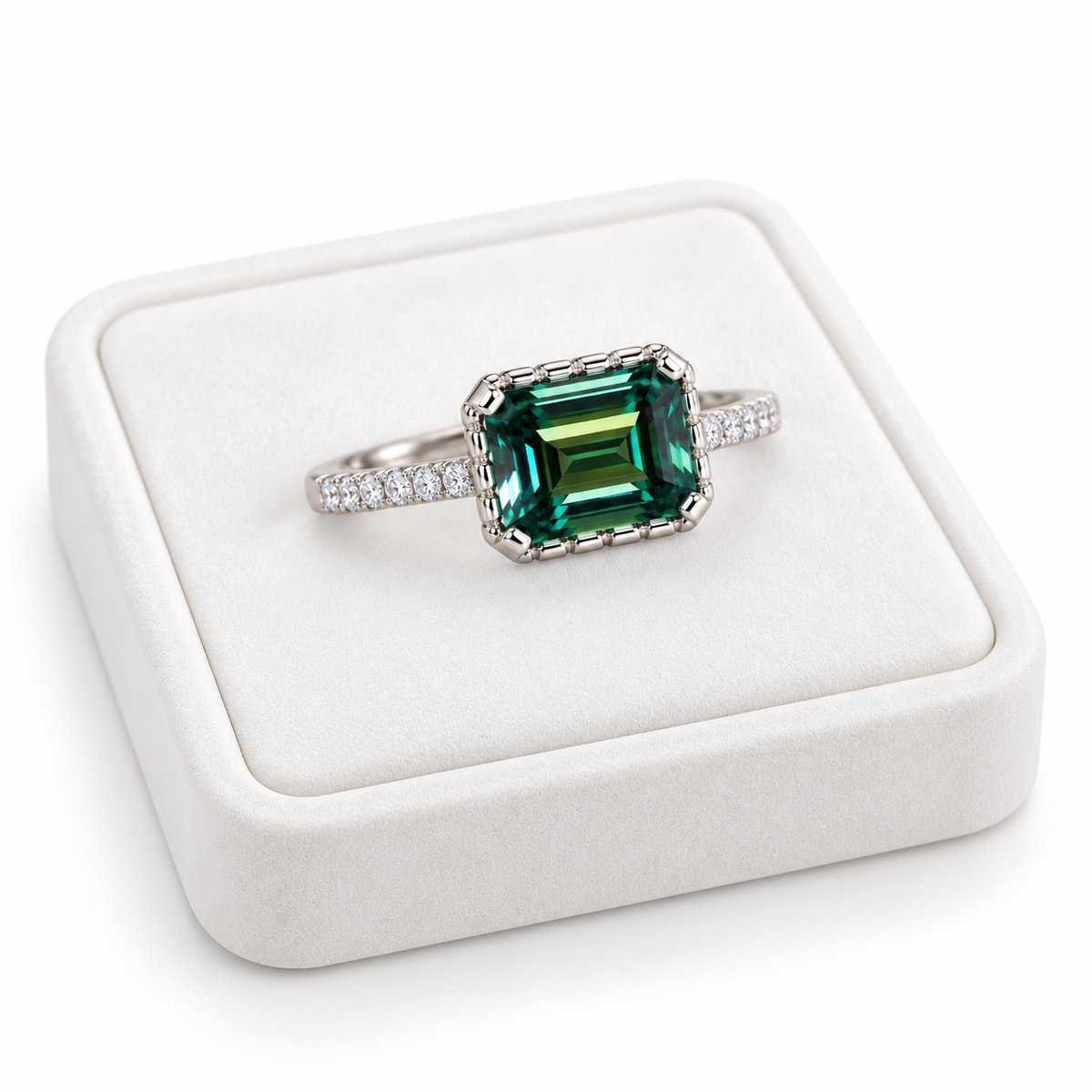 Angled photograph of a 2 carat lab-created green moissanite ring resting on a white velvet jewelry tray, showcasing the elongated 6x8mm emerald-cut gemstone, teal-green reflections, and Platinum Plated sterling silver setting.