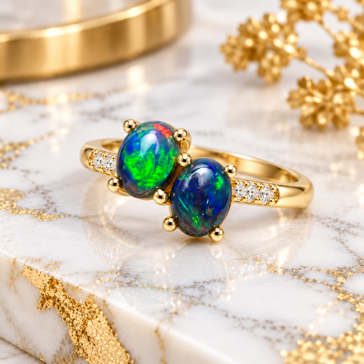 Angled lifestyle photograph of a black fire opal two-stone ring resting on white marble, showcasing the offset oval gemstones, sparkling cubic zirconia accents, and polished gold-colored band.