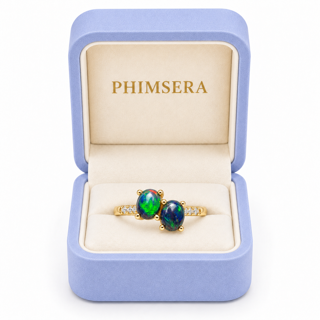 Black fire opal two-stone ring featuring two 4x5mm oval gemstones displayed inside a periwinkle Phimsera jewelry box with cream velvet interior and gold PHIMSERA lettering.
