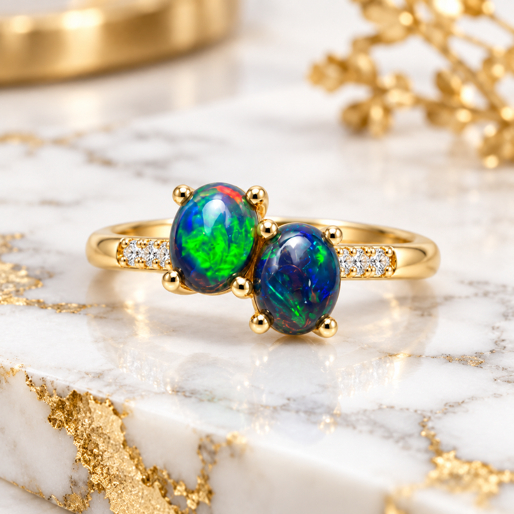 Black fire opal two-stone ring featuring two 4x5mm oval gemstones displayed on elegant white marble with subtle gray veining, highlighting the vivid blue-green opals and Gold Plated sterling silver setting.