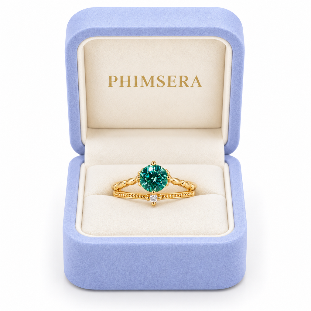 6.5mm round brilliant green moissanite ring displayed inside a periwinkle Phimsera jewelry gift box with an ivory interior and gold PHIMSERA lettering, showcasing the vibrant teal-green gemstone, elegant Gold Plated sterling silver band, and delicate cubic zirconia accent against a white background.