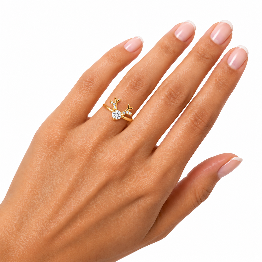 A gold ring with a small clear gemstone set in a deer-head motif, shown worn on a hand with manicured nails against a white background.