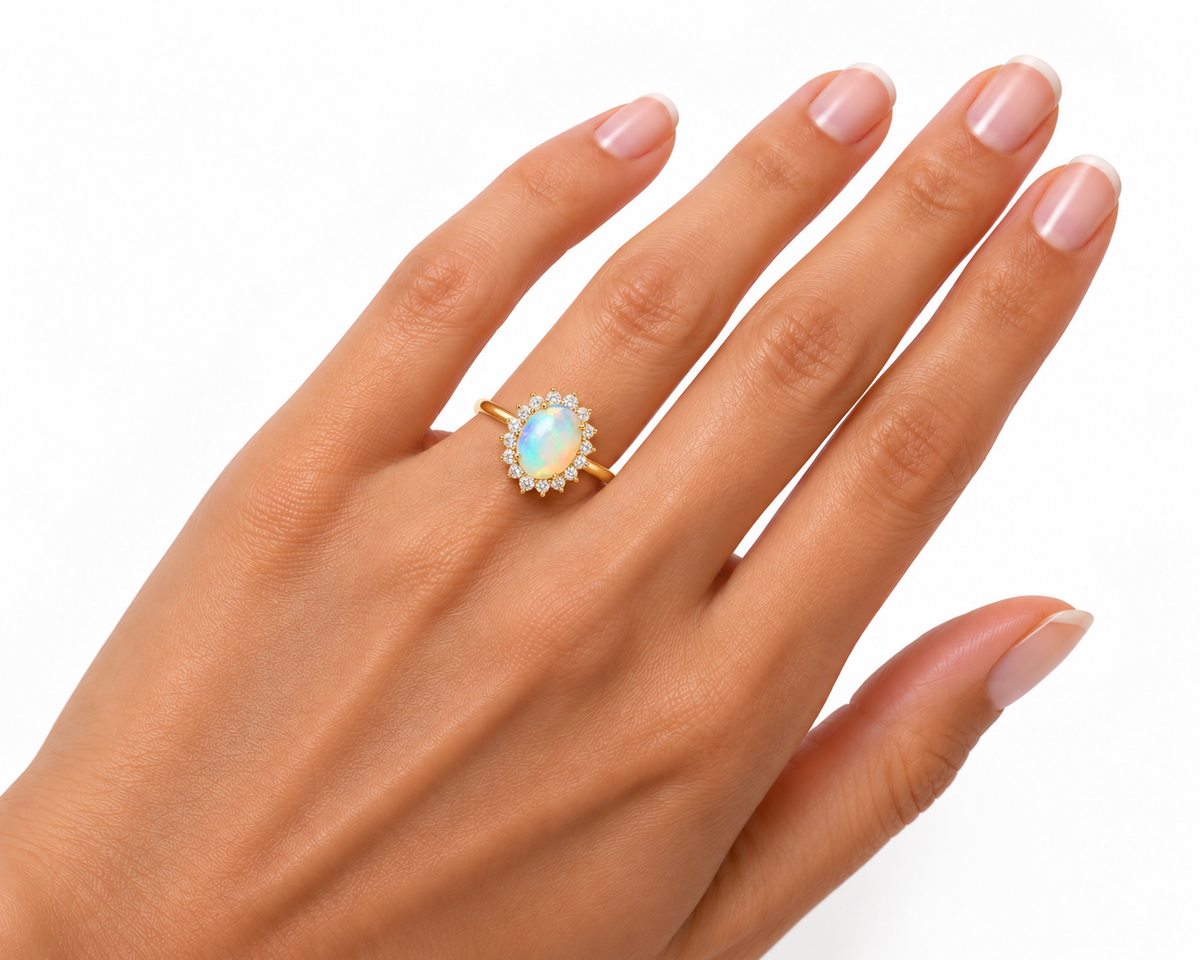 A gold ring featuring an oval iridescent opal stone surrounded by small clear stones, shown worn on a hand against a white background.