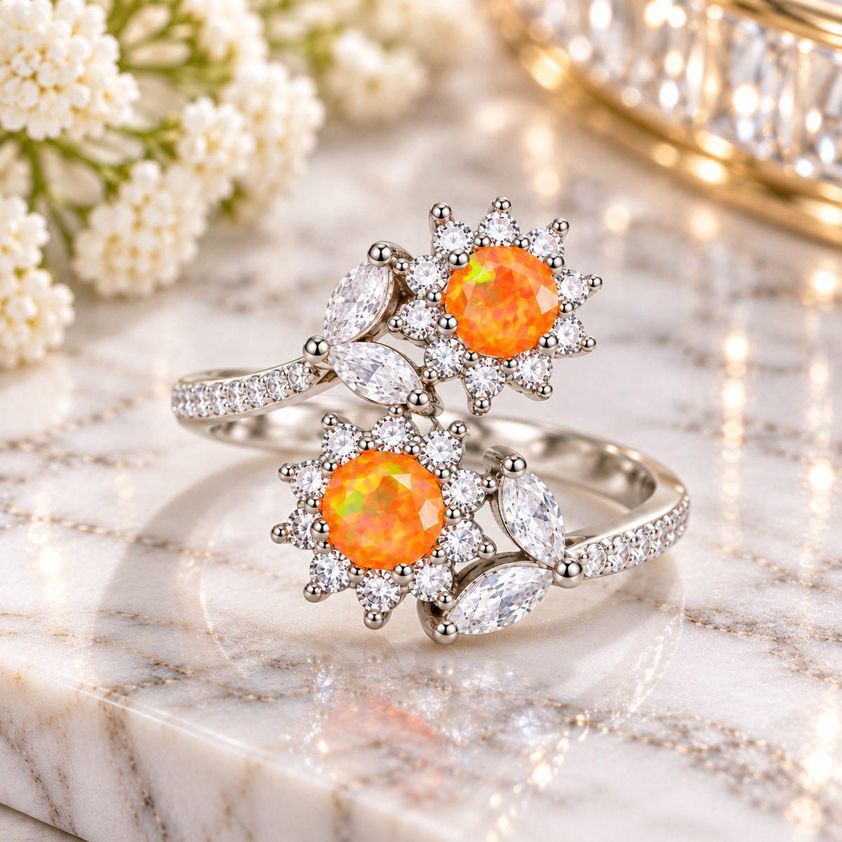 A pair of silver rings with vibrant orange opal gemstones and clear marquise and round crystals, displayed on a white marble surface with white flowers in the background.