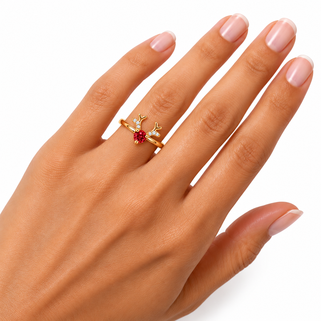 A gold ring with a small red heart-shaped gemstone and antler-like details, shown worn on a hand with light pink manicured nails against a white background.