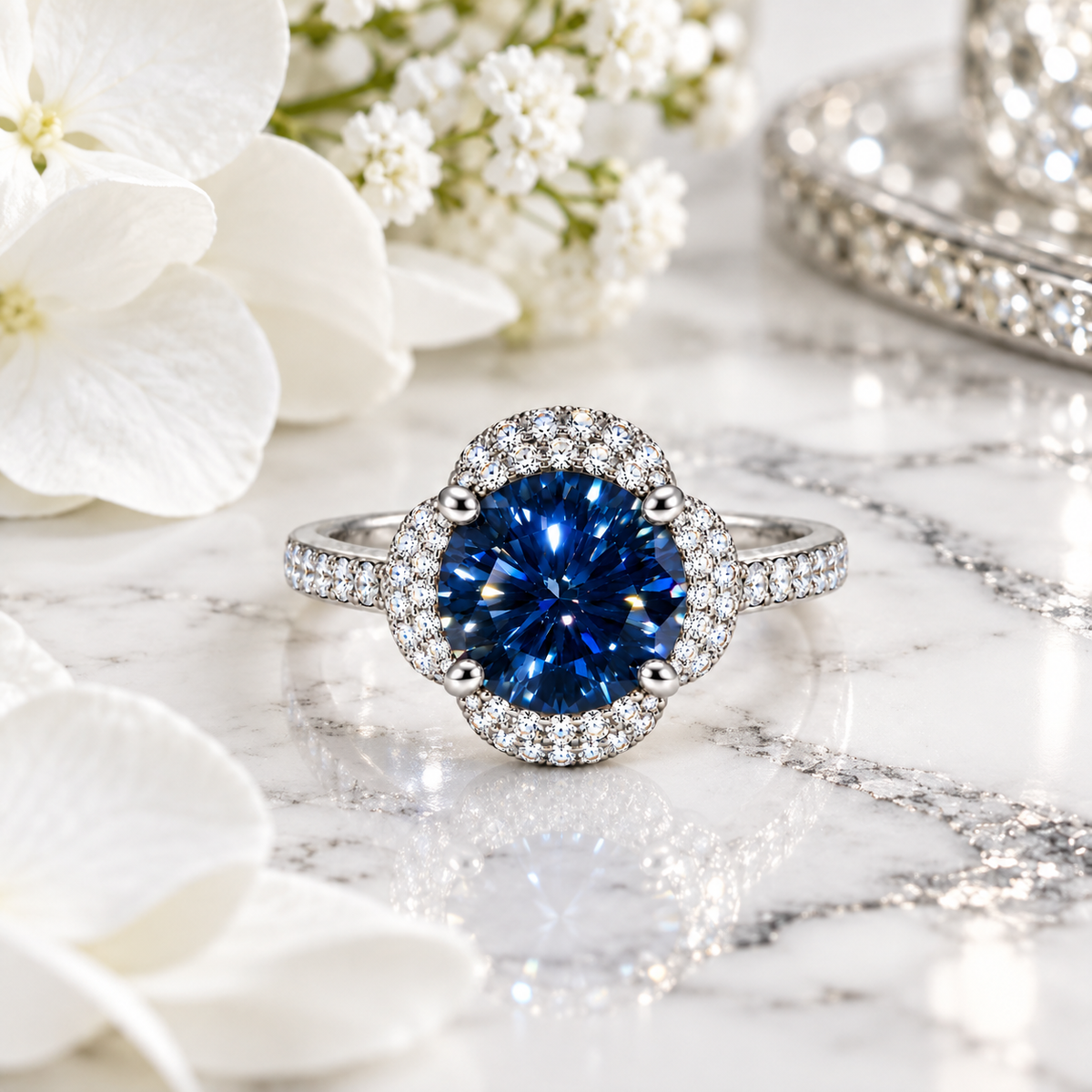 Phimsera 2 carat deep blue moissanite floral halo ring displayed on polished white marble with silver-gray veining, surrounded by pure white flowers and reflective silver accents.