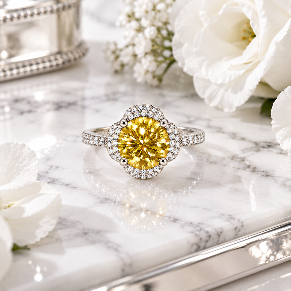 Phimsera 2 carat yellow moissanite floral halo ring displayed on polished white marble with silver-gray veining, surrounded by pure white flowers and reflective silver accents.