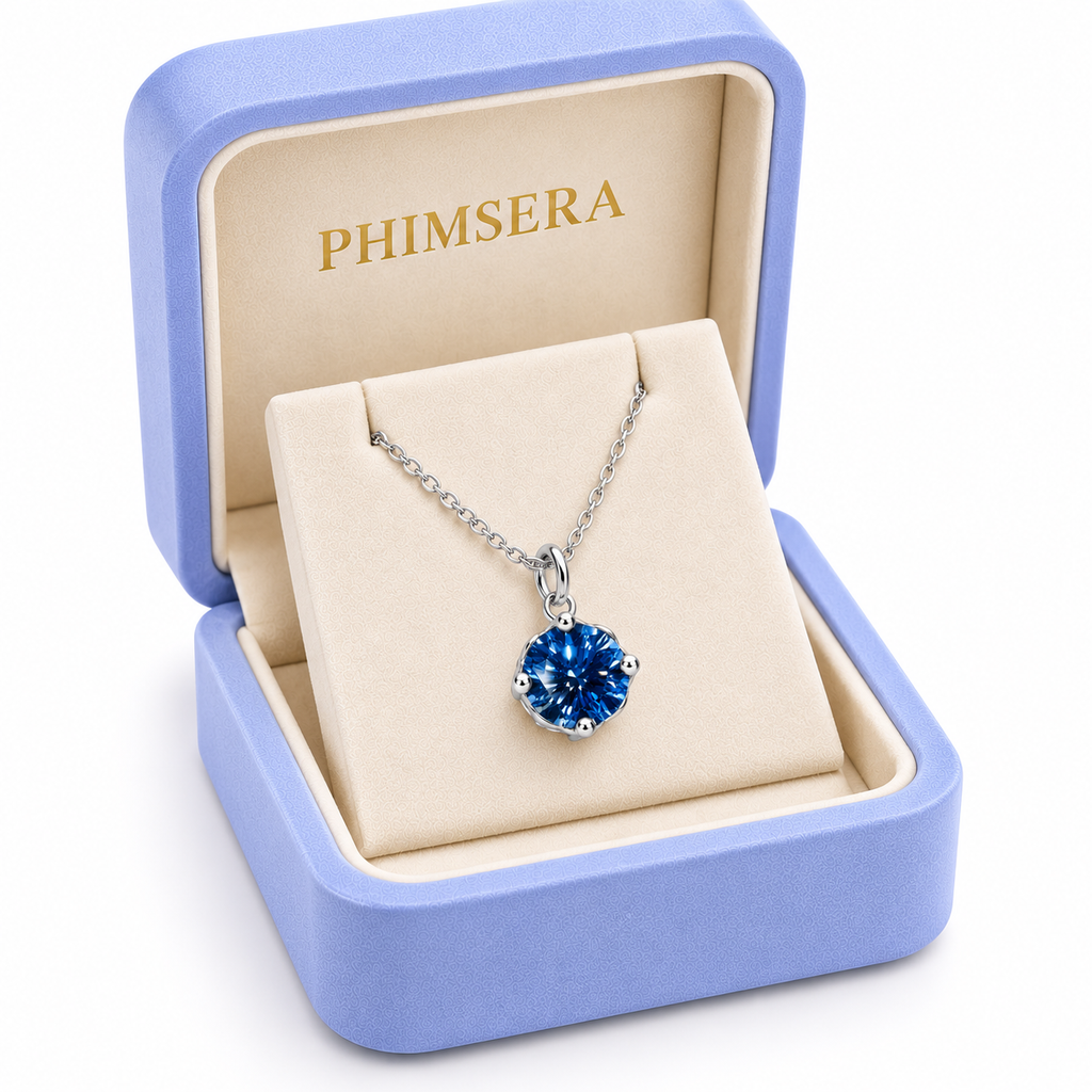 Angled view of a periwinkle Phimsera jewelry gift box containing an 8mm royal blue moissanite solitaire pendant necklace with a delicate silver chain, ivory jewelry insert, and gold Phimsera branding.