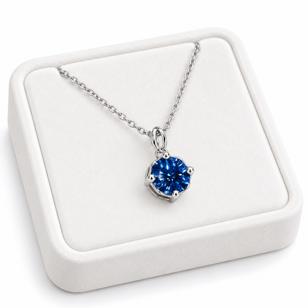 Angled white velvet jewelry tray displaying an 8mm round brilliant deep blue moissanite pendant necklace with a four-prong sterling silver setting, delicate oval-link chain, and clean white background.