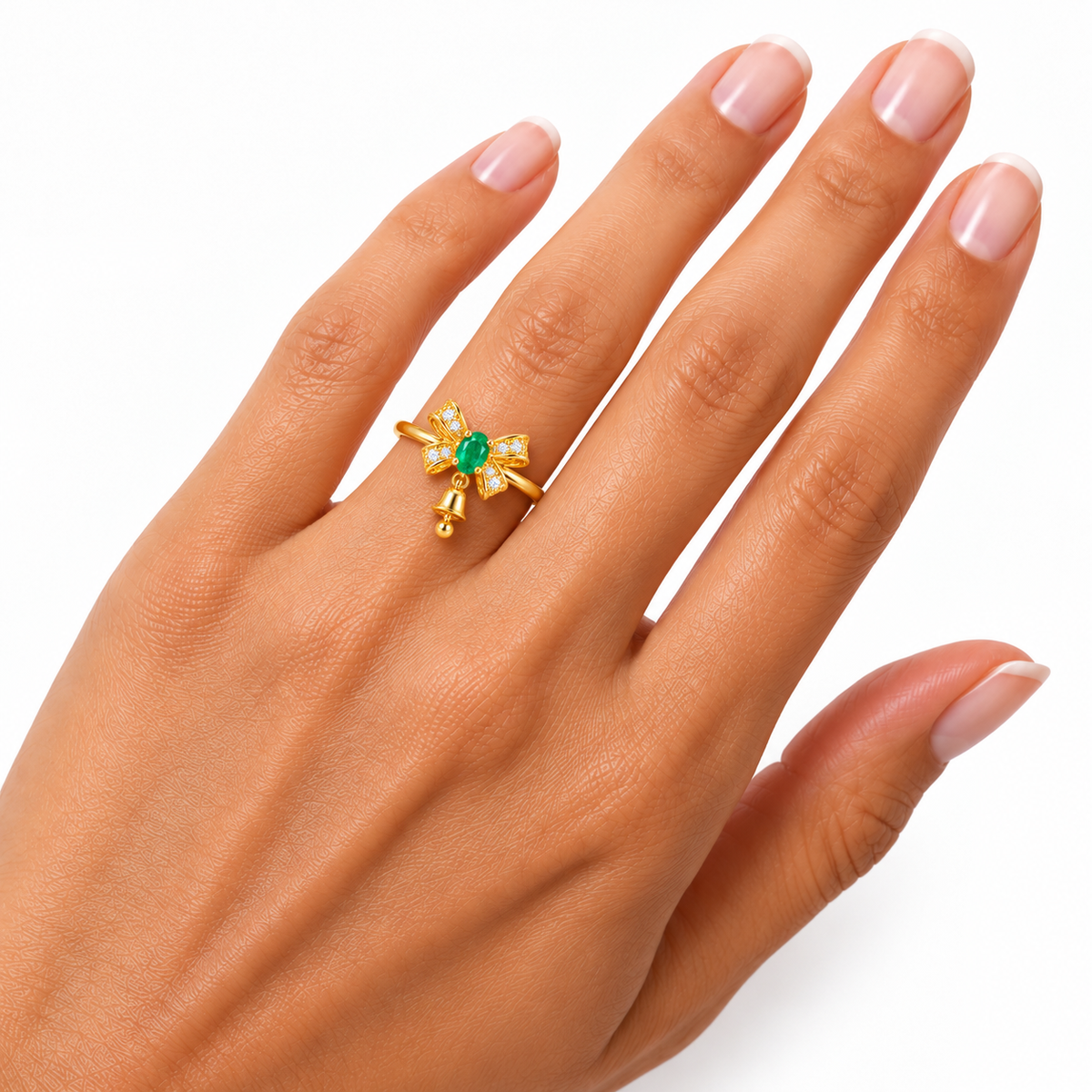Petite natural emerald Christmas bow ring worn on a woman's ring finger, showing a 3x4mm oval green emerald, delicate gold band, sparkling CZ bow and tiny dangling bell against white.
