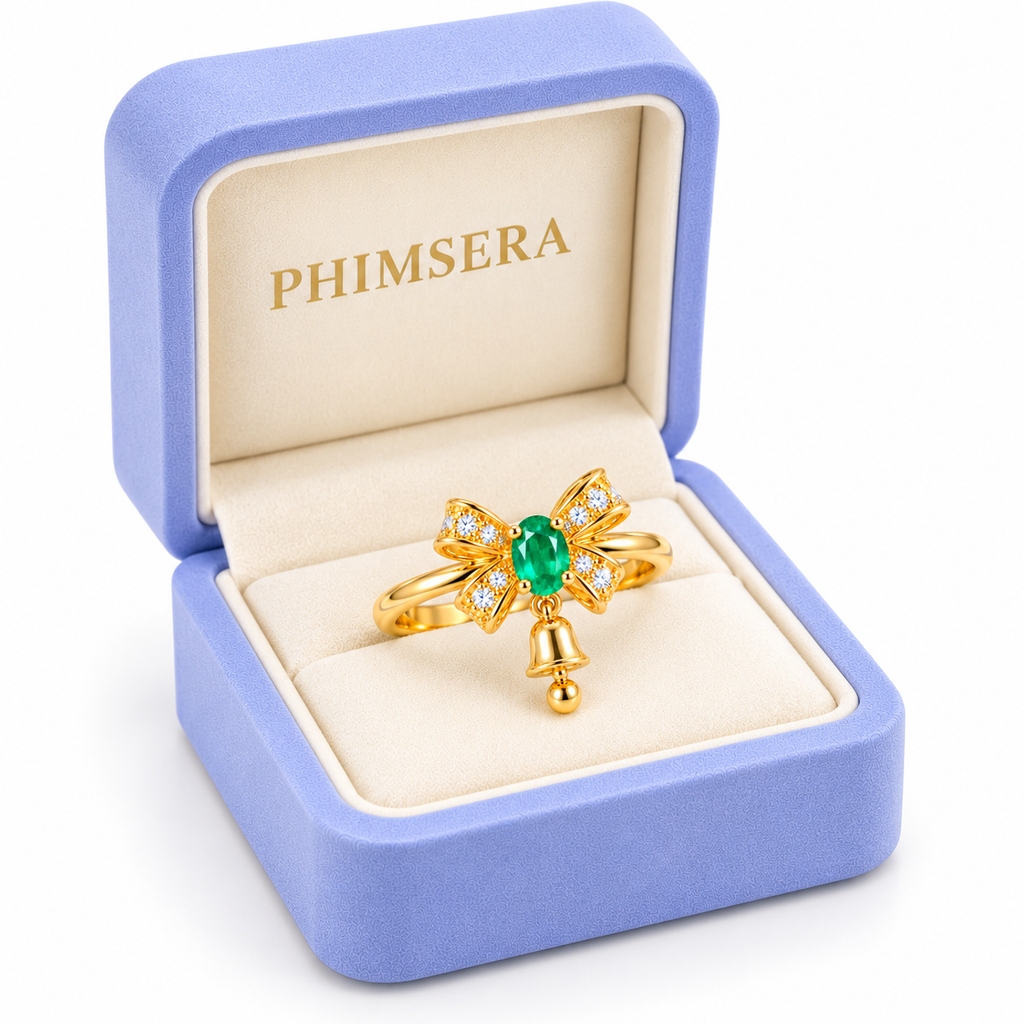 Angled view of a natural emerald bow and bell ring displayed inside a periwinkle Phimsera jewelry box, featuring a soft ivory interior, gold branding and a white studio background.