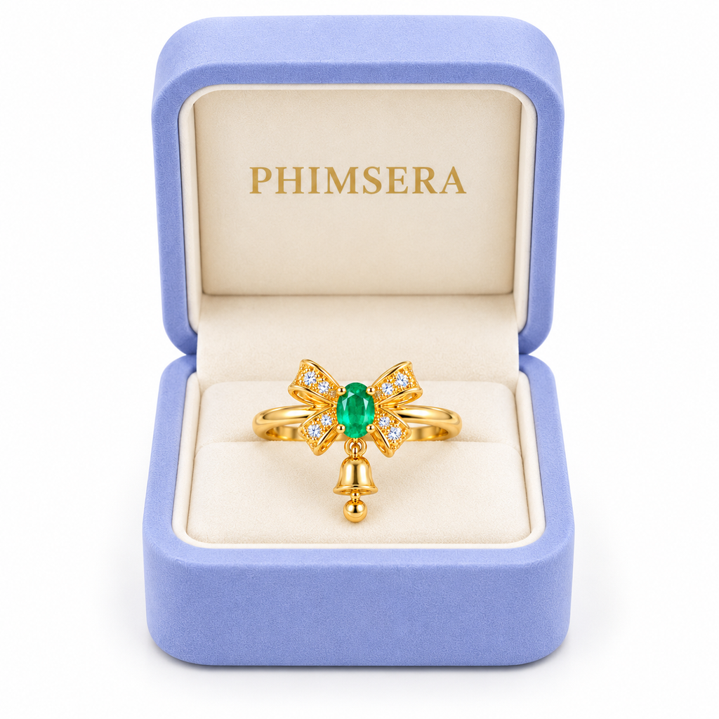 Natural emerald Christmas bow ring with a petite dangling gold bell displayed inside an open periwinkle Phimsera jewelry gift box with an ivory interior and gold PHIMSERA logo against a white background.