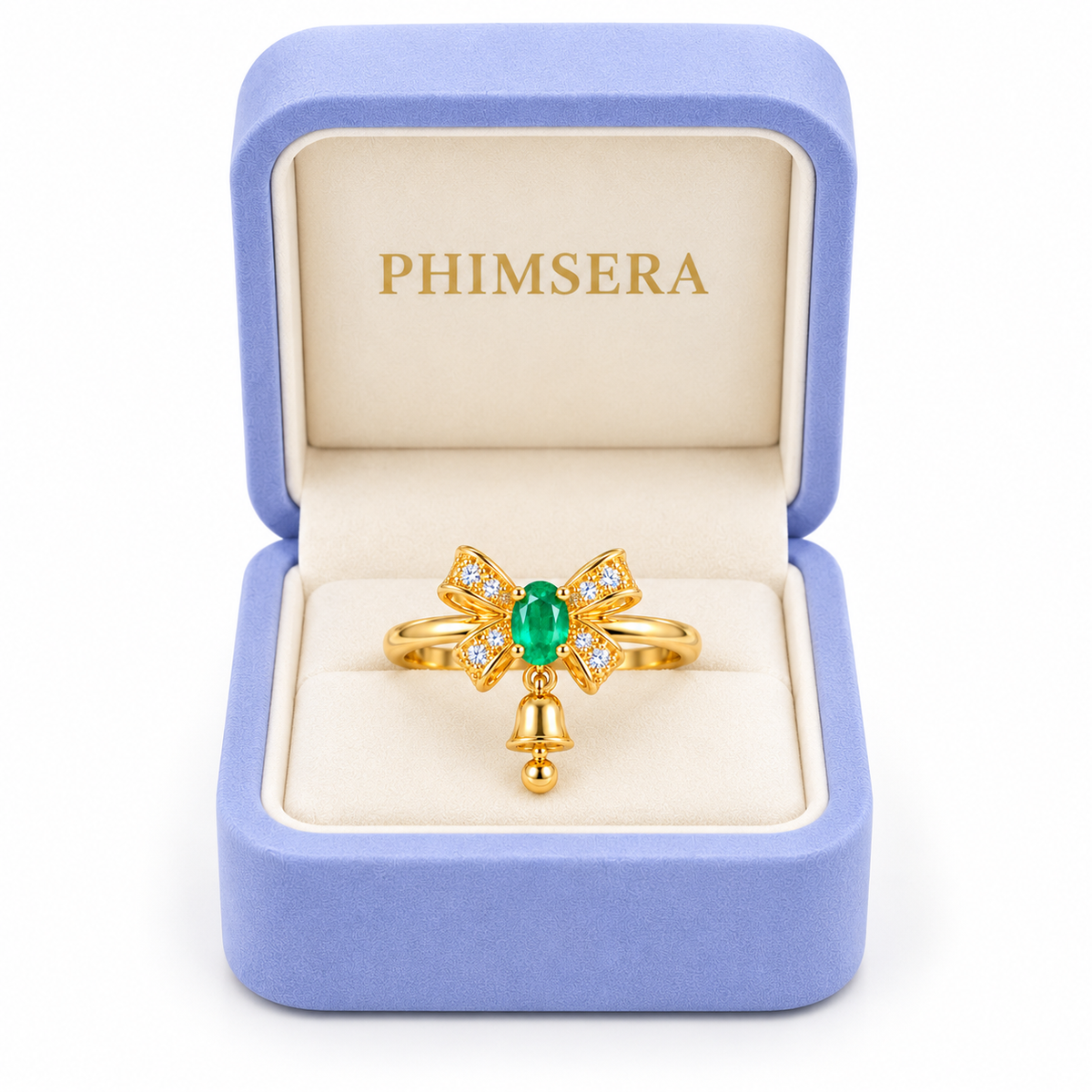 Natural emerald Christmas bow ring with a petite dangling gold bell displayed inside an open periwinkle Phimsera jewelry gift box with an ivory interior and gold PHIMSERA logo against a white background.