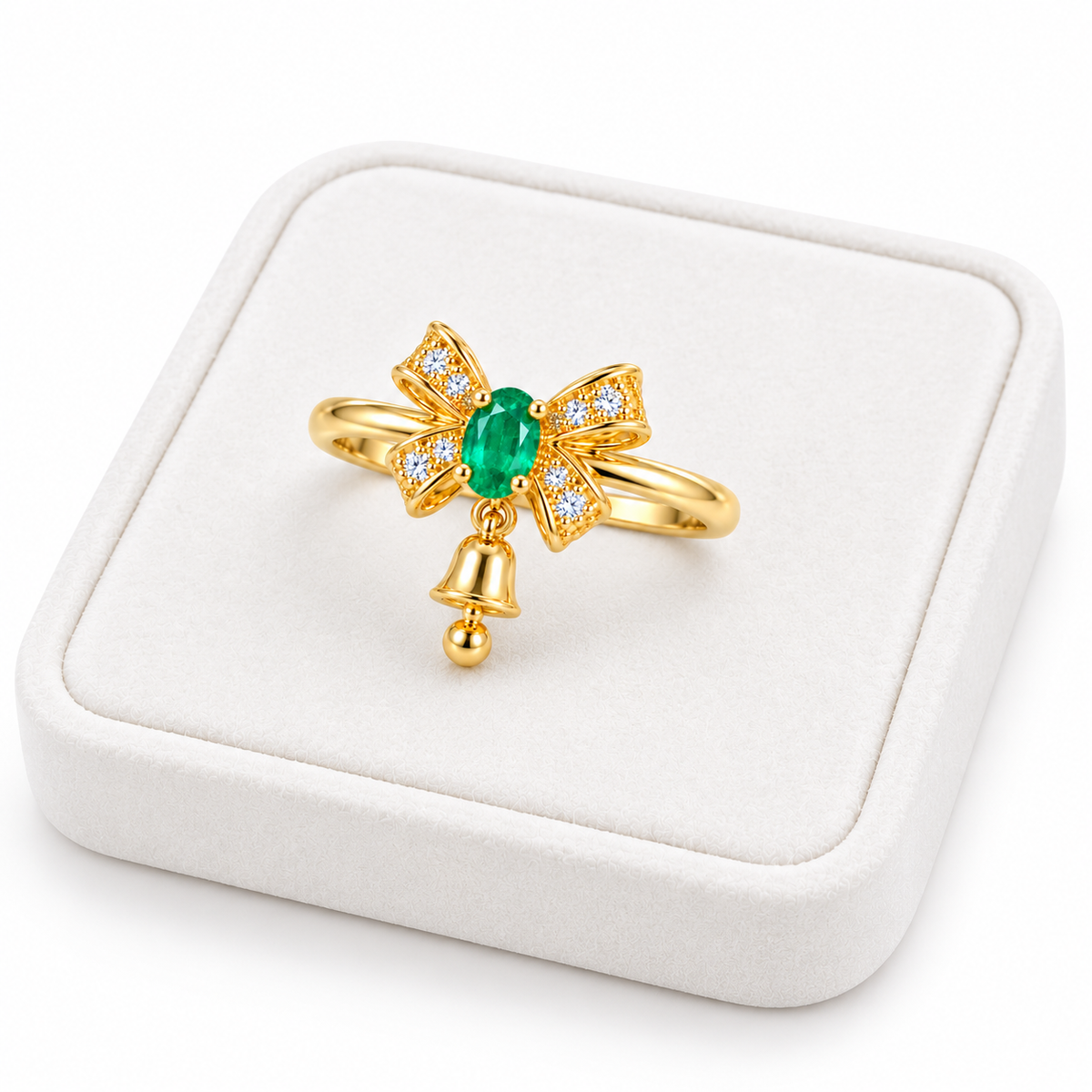 Angled white velvet tray presentation of a gold-plated sterling silver emerald bow ring showing its vivid green gemstone, sparkling ribbon design and petite hanging Christmas bell.