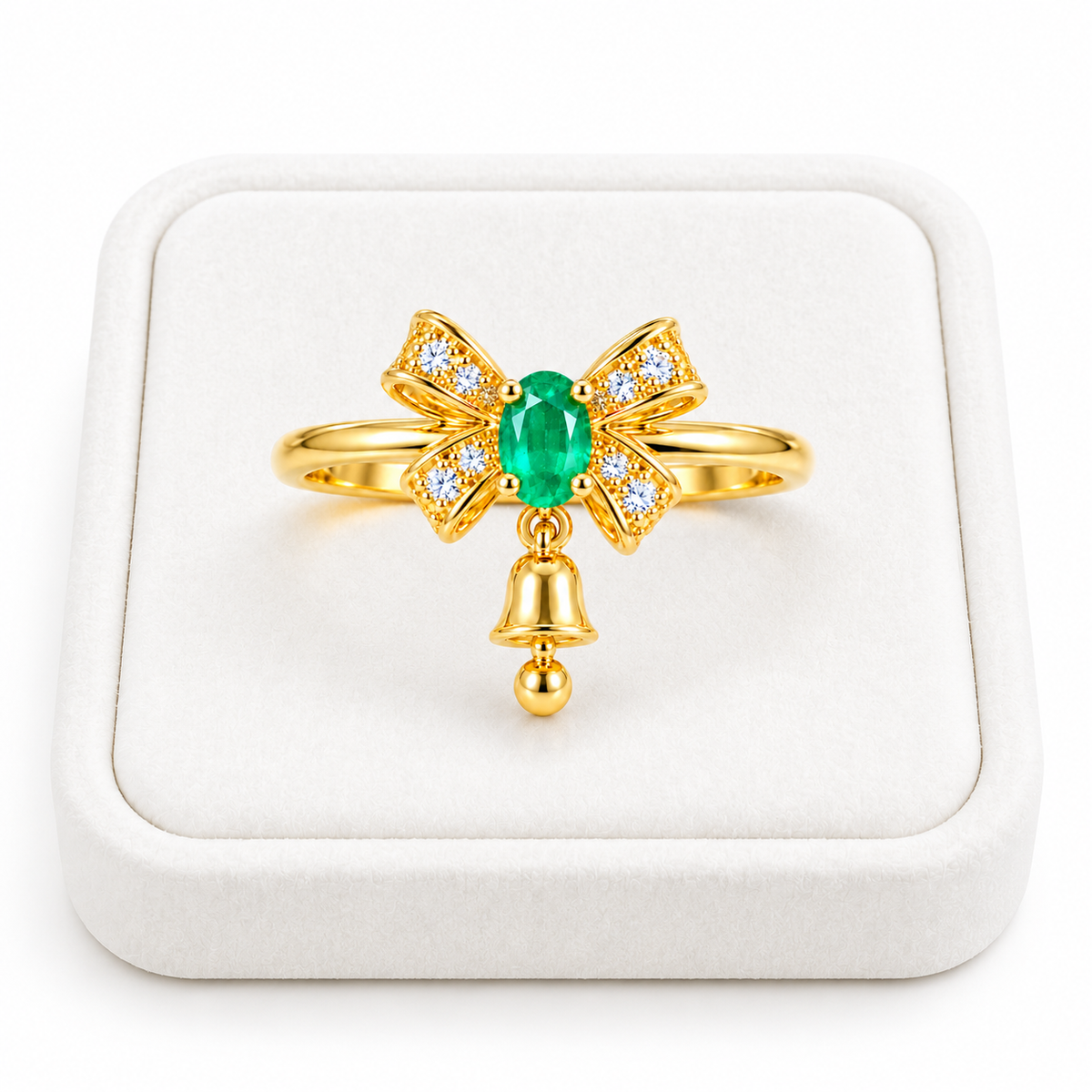 Natural 3x4mm oval emerald Christmas bow ring with white CZ accents and a dangling gold bell displayed on a premium white velvet jewelry tray against a pure white background.