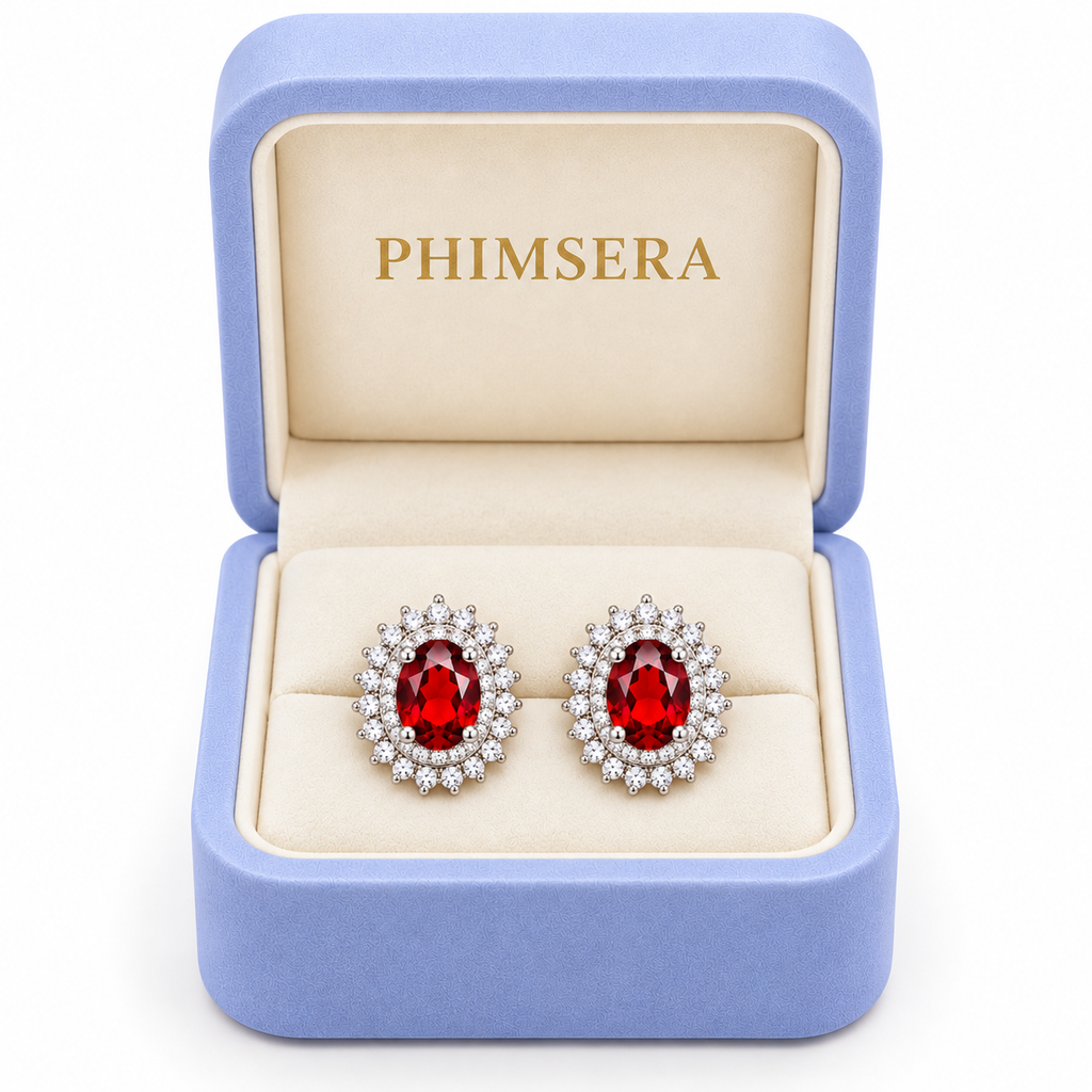 Natural red garnet double halo stud earrings displayed inside an open periwinkle Phimsera jewelry gift box with an ivory interior and gold Phimsera logo, featuring 5x7mm oval crimson gemstones and sparkling cubic zirconia halos against a white background.