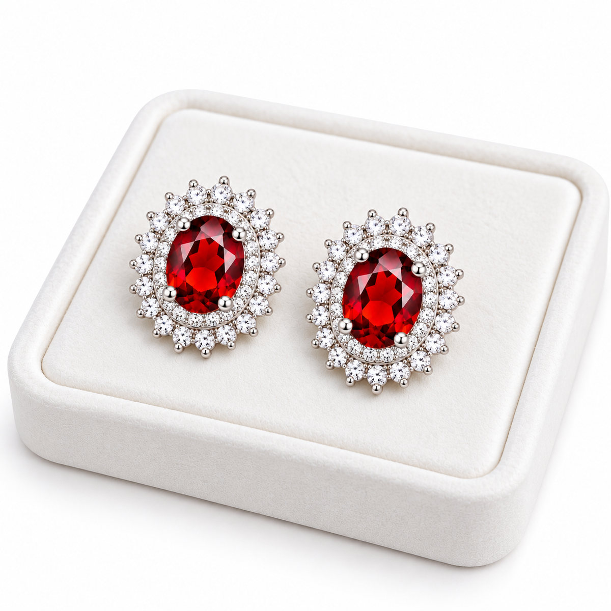 Angled white velvet jewelry tray displaying matching natural red garnet double halo stud earrings with 5x7mm oval gemstones, polished sterling silver four-prong settings, and brilliant clear cubic zirconia accents on a pure white background.