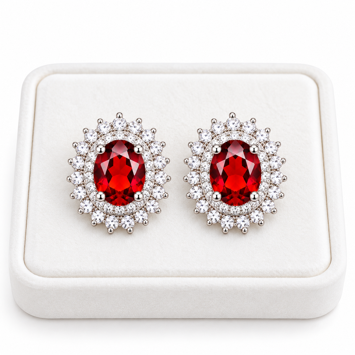 Pair of natural 5x7mm red garnet double halo stud earrings displayed side by side on a premium white velvet jewelry tray, featuring crimson-red oval gemstones, sparkling cubic zirconia sunburst halos, and polished sterling silver settings against a white background.