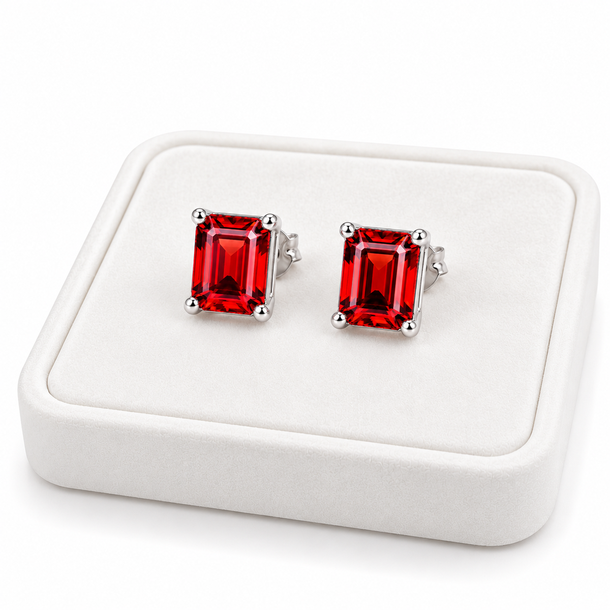 Angled white velvet jewelry tray displaying matching natural red garnet emerald cut stud earrings with 6x8mm gemstones, polished sterling silver settings, and brilliant crimson-red reflections against a clean white background.