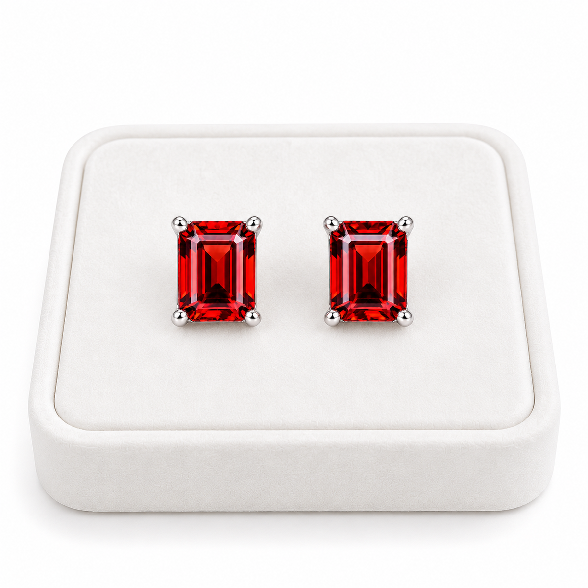 Pair of natural 6x8mm red garnet emerald cut stud earrings displayed upright on a premium white velvet jewelry tray, featuring crimson-red gemstones and polished sterling silver four-prong settings against a pure white background.