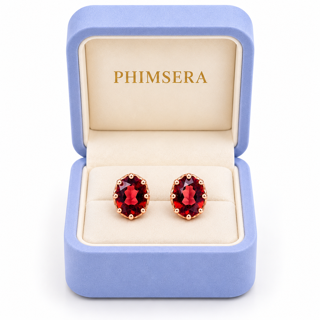 Natural 6x8mm red garnet oval stud earrings displayed inside an open periwinkle Phimsera jewelry gift box with an ivory interior, gold Phimsera logo, and pure white background.