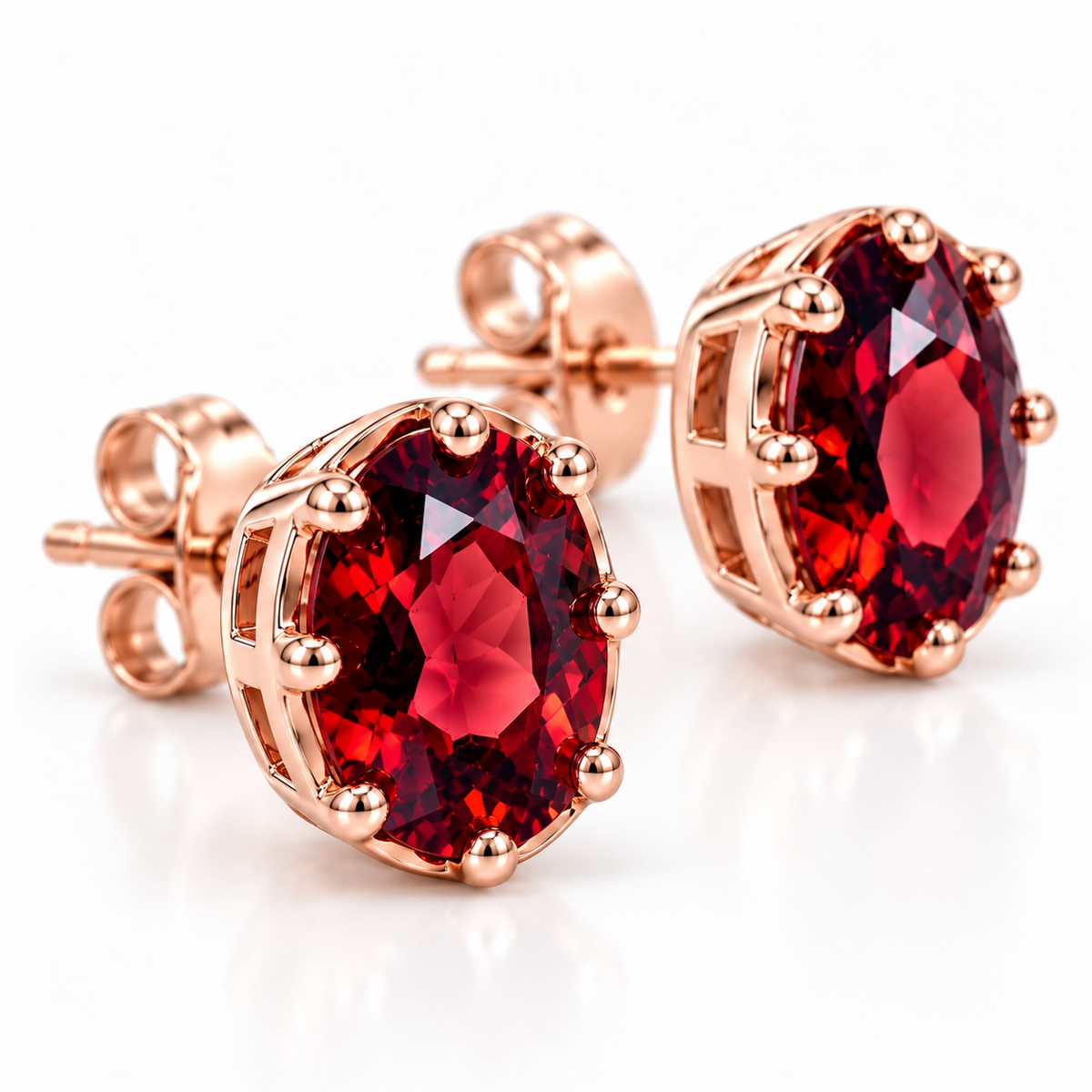 Angled macro photograph of natural 6x8mm oval red garnet stud earrings highlighting rich crimson-red facets, deep burgundy reflections, polished rose gold plated prongs, and detailed open-gallery settings.