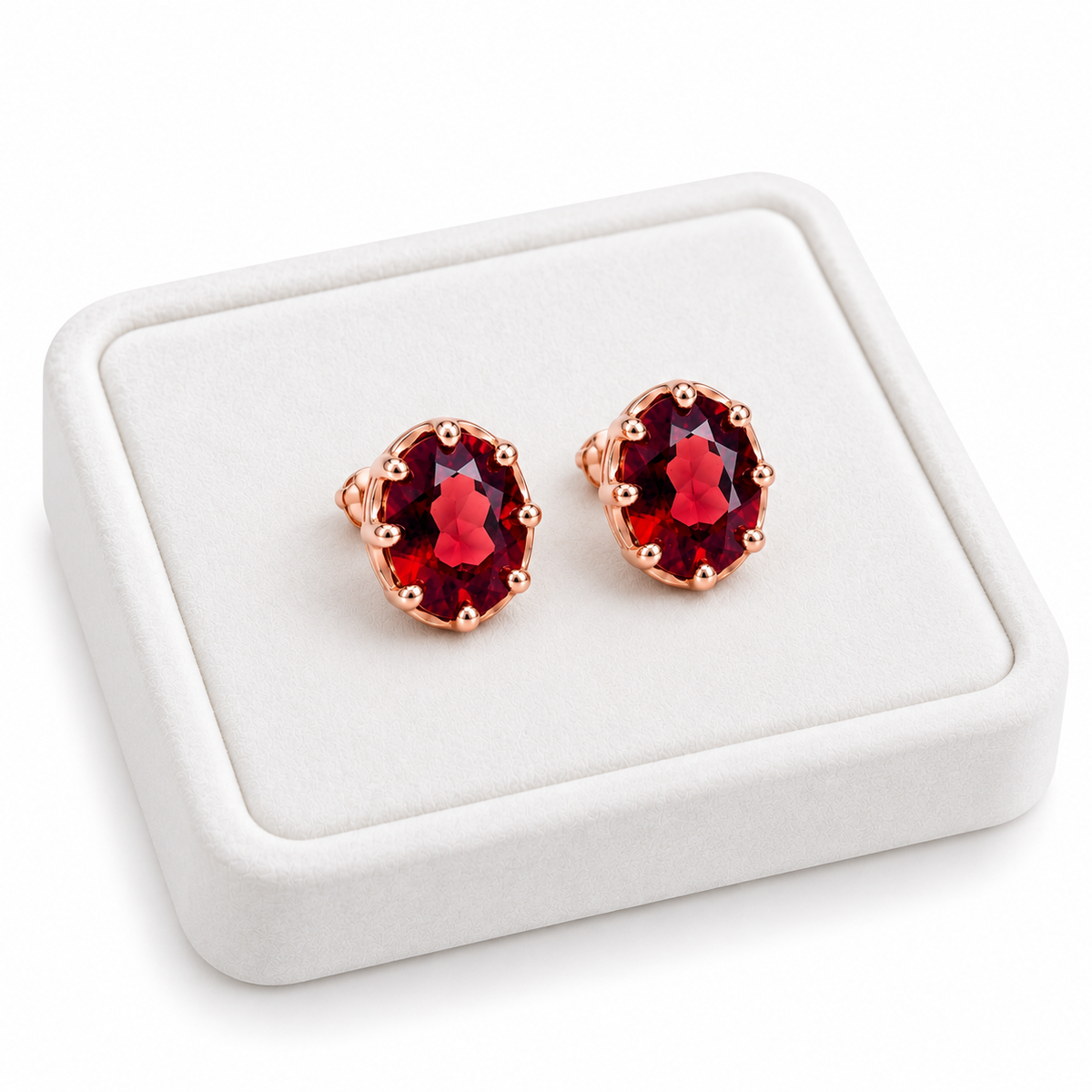 Angled white velvet jewelry tray displaying matching natural red garnet oval stud earrings with 6x8mm gemstones, polished rose gold plated settings, and brilliant crimson-red reflections on a clean white background.