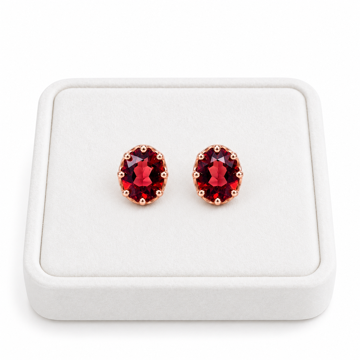 Pair of natural 6x8mm red garnet oval stud earrings displayed upright on a premium white velvet jewelry tray, featuring rich crimson gemstones and polished rose gold plated sterling silver settings against a pure white background.