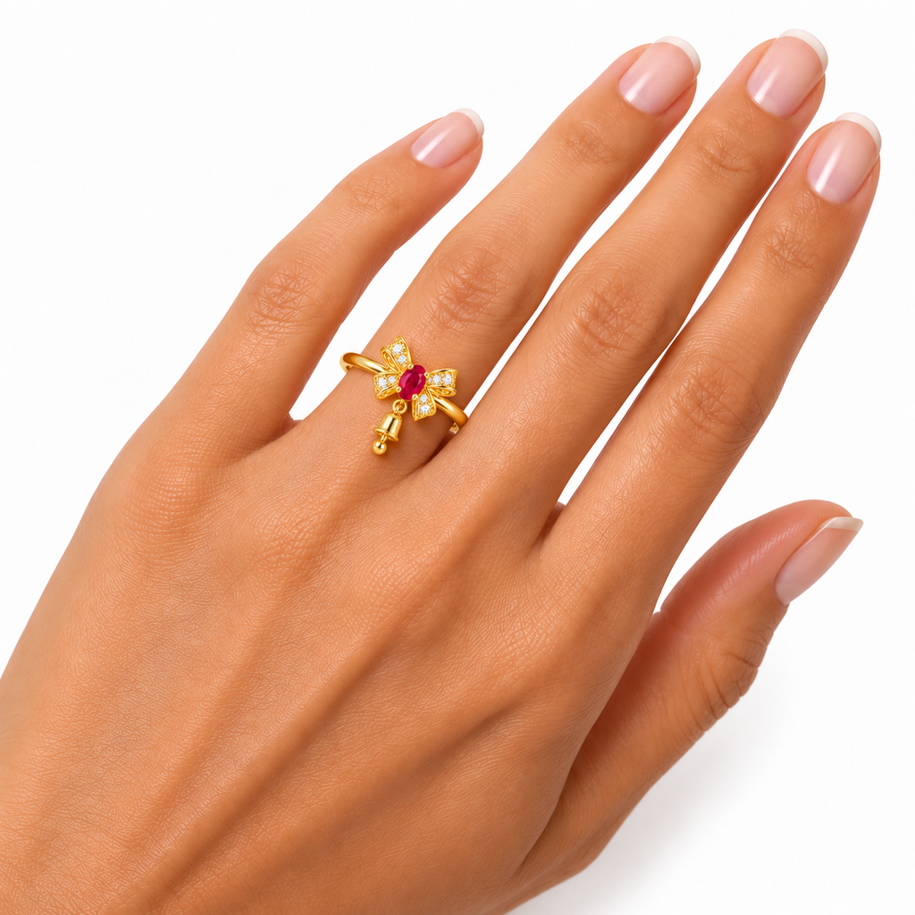 Petite natural ruby Christmas bow ring worn on a woman's ring finger, showing a true-to-size 3x4mm oval red ruby, delicate gold band, sparkling CZ bow and tiny dangling bell against white.