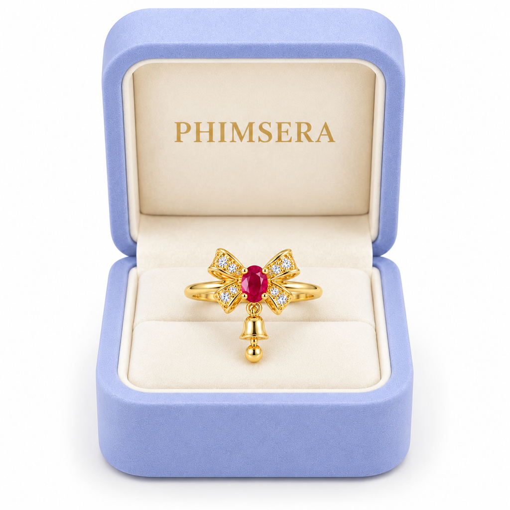 Natural ruby Christmas bow ring with a petite dangling gold bell displayed inside an open periwinkle Phimsera jewelry gift box with an ivory interior and gold PHIMSERA logo against a white background.