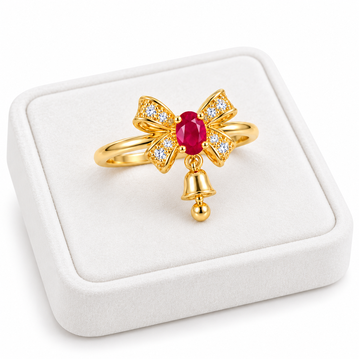 Angled white velvet tray presentation of a gold-plated sterling silver ruby bow ring showing its vivid red gemstone, sparkling ribbon design and petite hanging Christmas bell.
