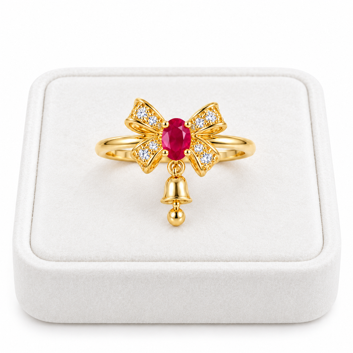Natural 3x4mm oval ruby Christmas bow ring with white CZ accents and a dangling gold bell displayed on a premium white velvet jewelry tray against a pure white background.