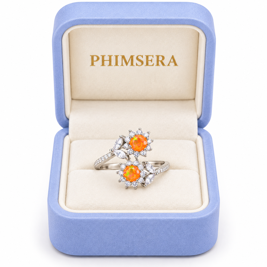 A pair of silver rings with orange gemstone clusters and clear stones, presented in an open light blue PHIMSERA jewelry box on a white background.